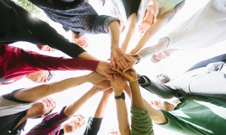 A group of people standing in a circle with their hands stacked together in the center, viewed from below.