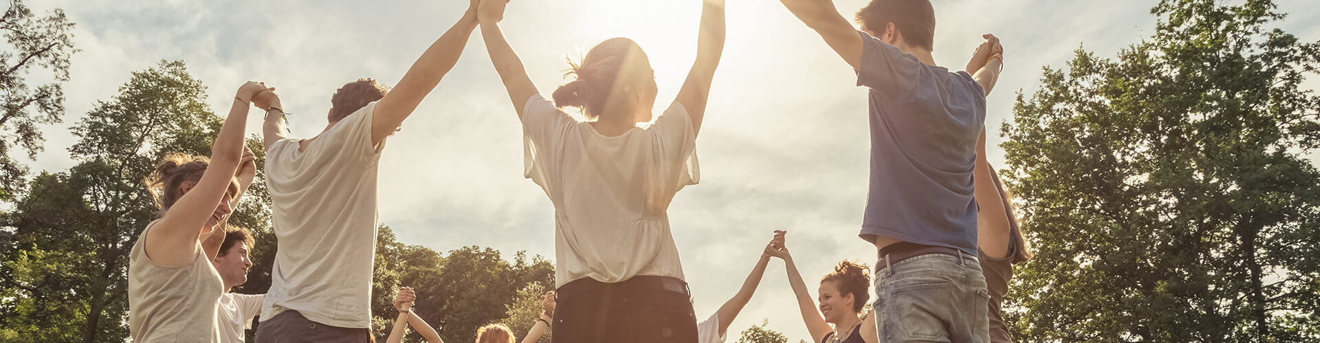 A group of people standing outdoors in a circle holding hands, with the sun shining in the background and trees surrounding them.