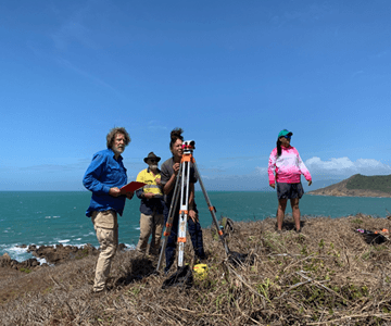 Four people stand on a grassy coastal hill with a tripod and equipment, overlooking the ocean under a clear blue sky.