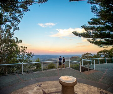Two people stand at a scenic lookout with a railing, trees, and a wide view of the landscape and sky at sunset. A round stone structure is in the foreground.