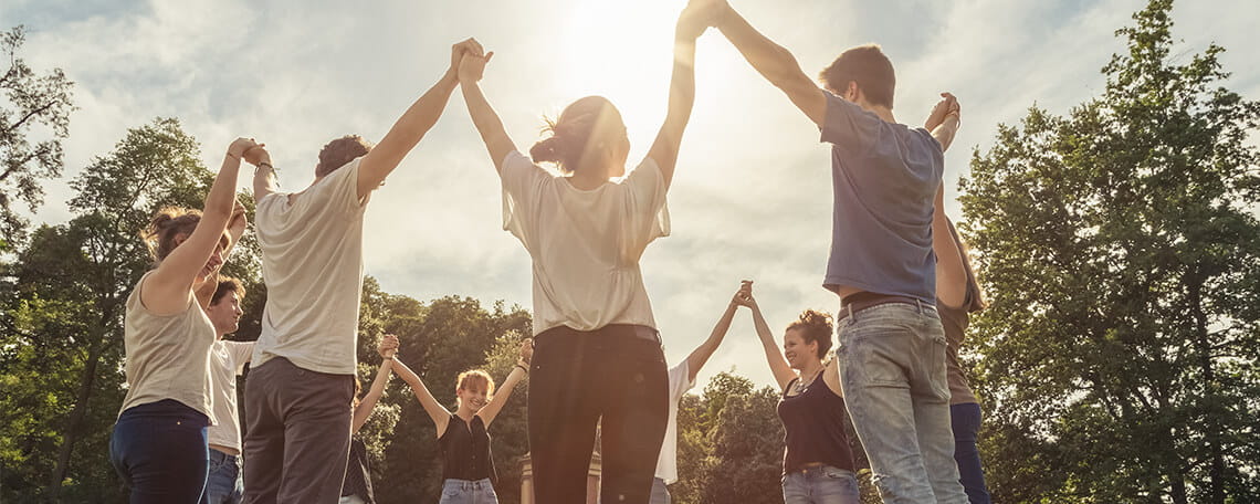 A group of people standing in a circle outdoors, holding hands and raising their arms, with sunlight shining through the trees.