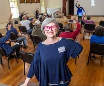 Woman with a "hello" sticker smiling at the camera in a room with people participating in a group activity.