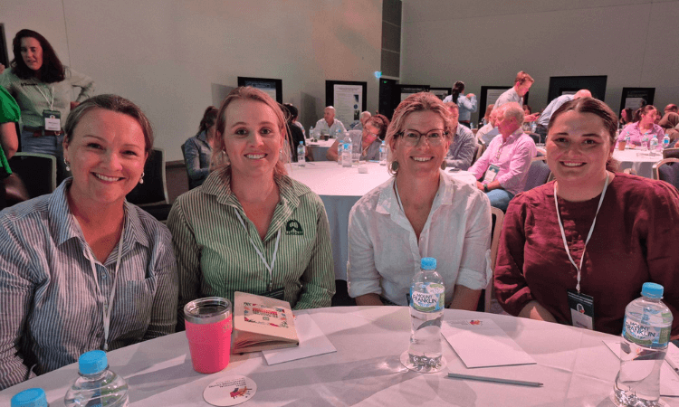 Four women sit at a round table with water bottles, a coffee cup, and notebooks, attending an indoor event with other participants in the background.