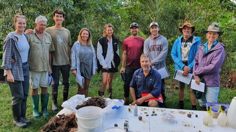 A group of ten people stands outdoors in front of trees, posing for a photo behind a table with soil samples and scientific tools.