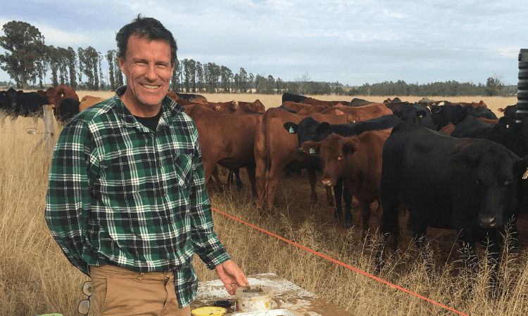 A man in a plaid shirt stands outdoors near a table with supplies, smiling in front of a herd of brown and black cattle in a grassy field.