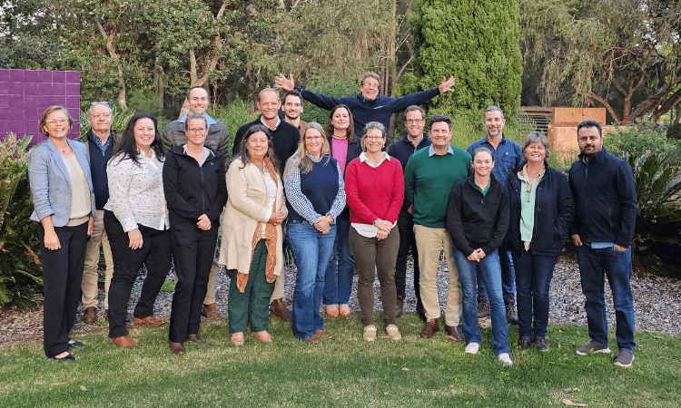 A group of seventeen adults stands and poses outdoors on grass, with trees and shrubs in the background. One person at the back raises their arms enthusiastically.