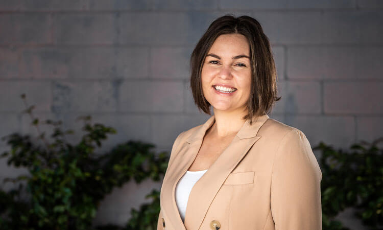 A smiling woman in a beige blazer standing in front of a gray brick wall with green plants in the background.