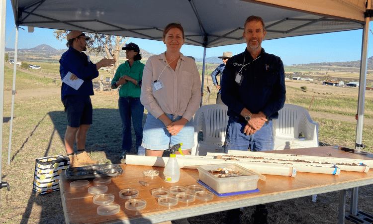 Two people, a woman and a man, stand behind a table with soil samples under a canopy at an outdoor event. Other individuals are seen in the background, engaging in conversation.