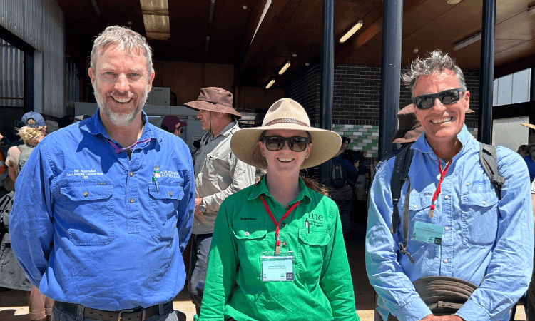 Three people wearing hats, name badges, and outdoor work shirts stand smiling in front of a building with others in the background.