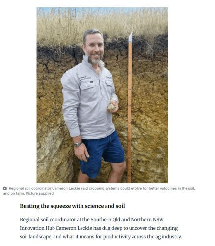 A man in outdoor clothing stands in a soil pit, holding a soil sample next to a measuring stick in a rural area.