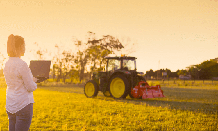 A person holding a laptop stands in a sunlit field, observing a tractor parked on the grass with trees and a fence in the background.