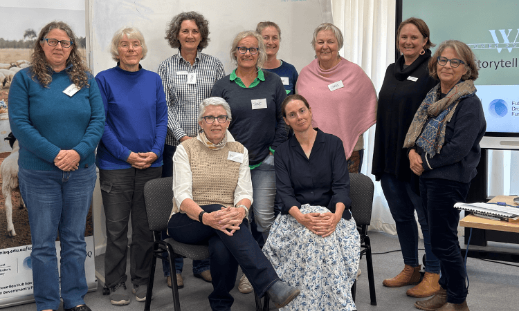 Ten women pose for a group photo indoors, with two seated at the front and eight standing behind them. Most are smiling and wearing name tags.
