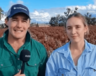 Two people stand in front of a field; the man on the left holds a microphone and wears a cap, while the woman on the right is dressed in a light blue shirt.