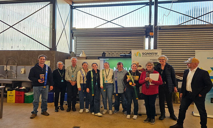 A group of people standing indoors in front of a wall, some holding certificates and wearing lanyards. There are banners and a table with objects in the background.