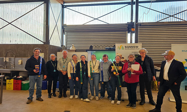 A group of people standing indoors in front of a wall, some holding certificates and wearing lanyards. There are banners and a table with objects in the background.
