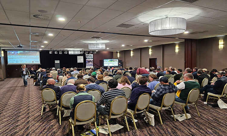 A conference room filled with attendees seated, facing a presenter and a projector screen at the front.