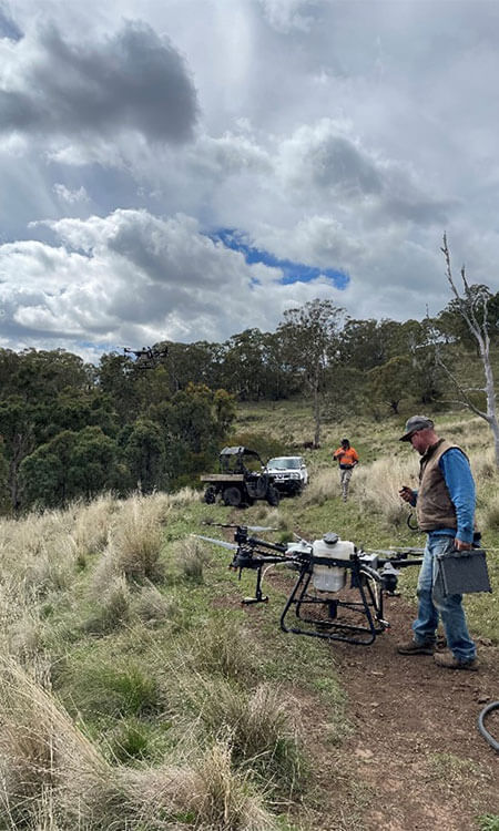 Two people with a large drone stand in a grassy field, with vehicles and cloudy sky in the background.