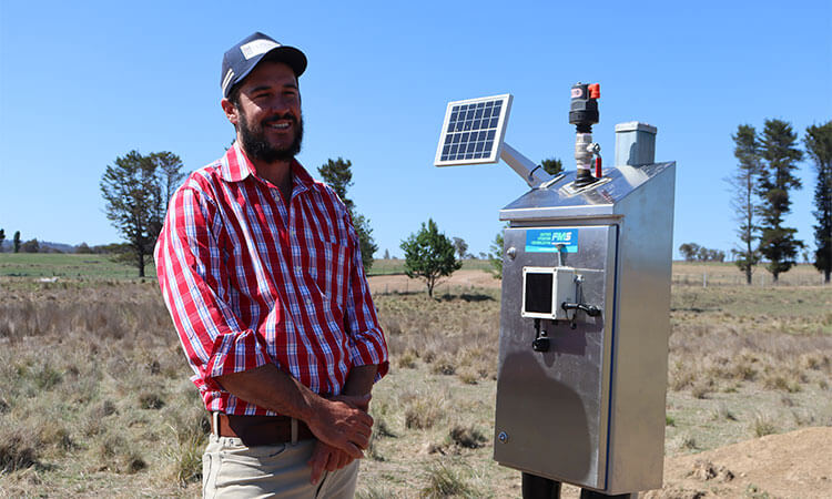 Man in a red plaid shirt stands next to a solar-powered device with a small screen in an open field under a clear blue sky.