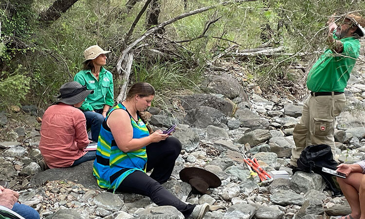 A group of people in outdoor clothing are sitting and standing on rocks in a wooded area, engaged in discussion or taking notes.