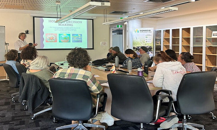 A presenter leads a discussion on holistic decision-making to a group seated around a conference table in a meeting room.