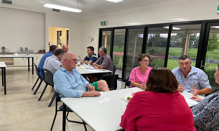 People seated at several tables in a bright room, engaged in discussion. Large windows show a view of greenery outside.