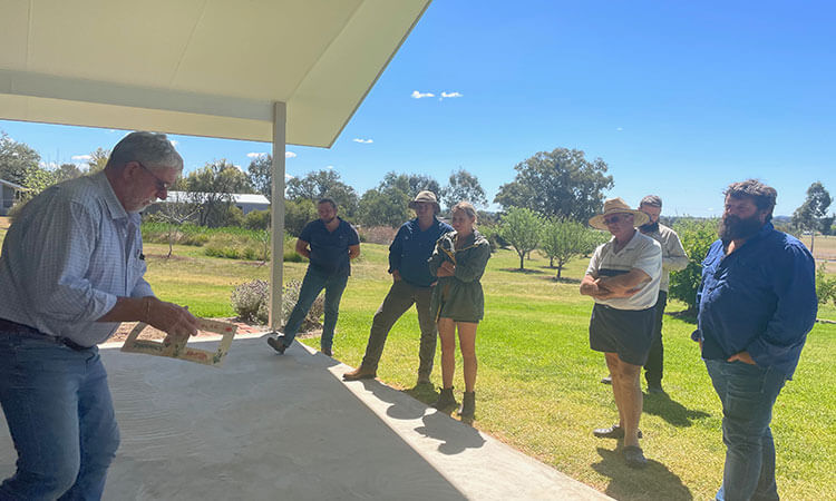 A group of people stands outdoors on a patio, listening to a person demonstrating or explaining something in a rural setting.