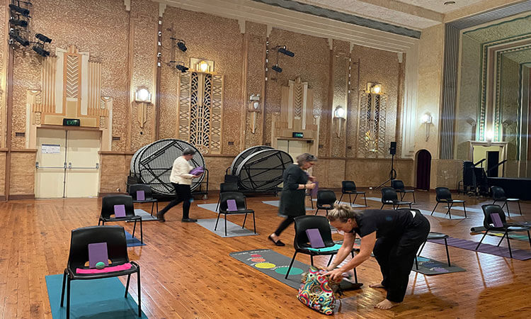 A room with wooden floors and beige walls, set up for a class or event. Chairs and yoga mats are arranged in a spacious layout, with a few people organizing the space.