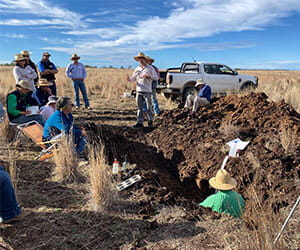 A group of people in a field observe a person standing near a large soil pit. A pickup truck is parked nearby under a partly cloudy sky.