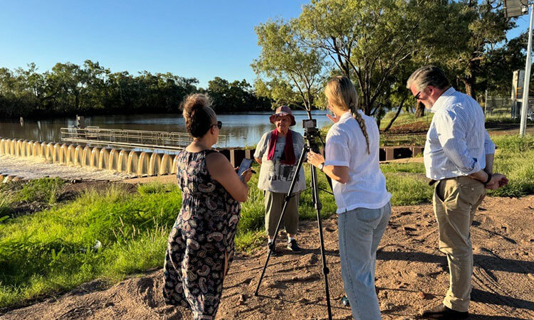 A person is being interviewed outdoors by three people with cameras near a river and bridge. Trees are in the background under a clear blue sky.