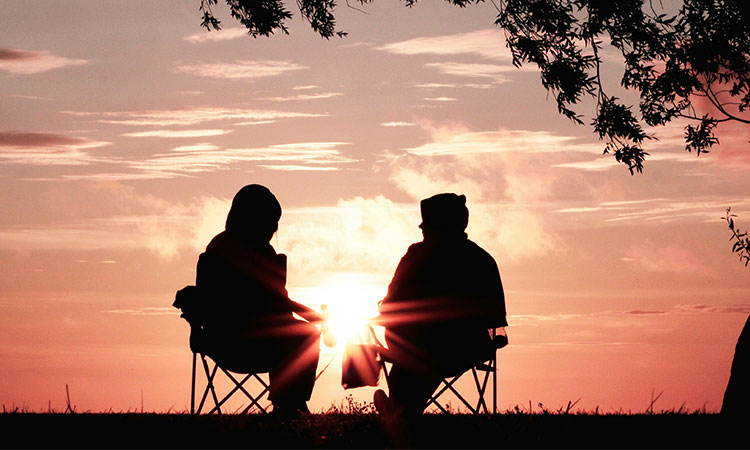 Two people seated, silhouetted against a sunset sky, with a tree on the right.