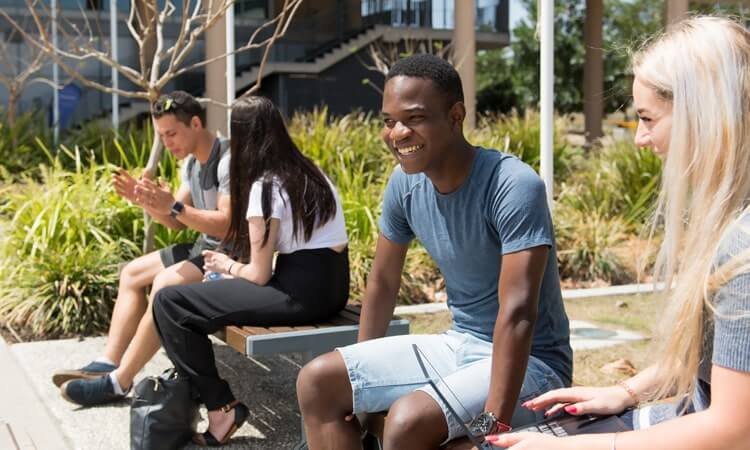 Four individuals casually sitting and enjoying the outdoors, with one man smiling towards the camera.