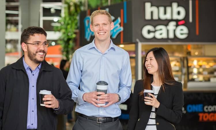 Three professionals holding coffee cups, walking and smiling in a corporate cafe setting.
