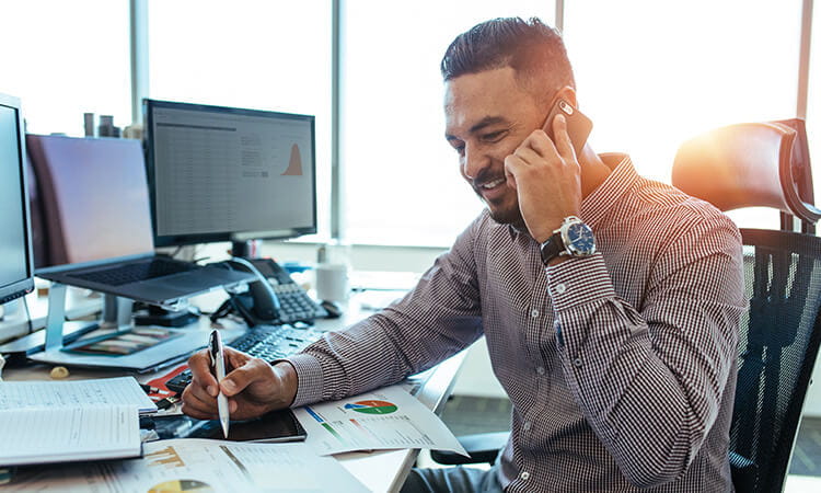 Man sitting at desk in front of computers, talking on phone and looking at printed information sheets. 