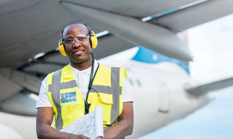 A male airport ground crew member wearing a reflective vest, hearing protection, and safety glasses stands in front of an airplane holding documents.