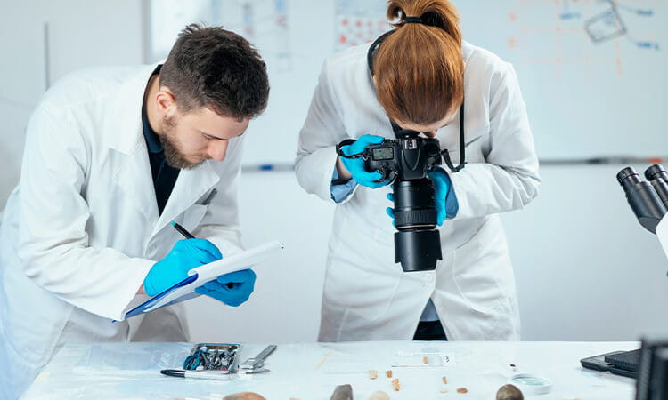 Two people in lab coats, one taking notes and the other using a camera to take photos of samples. 