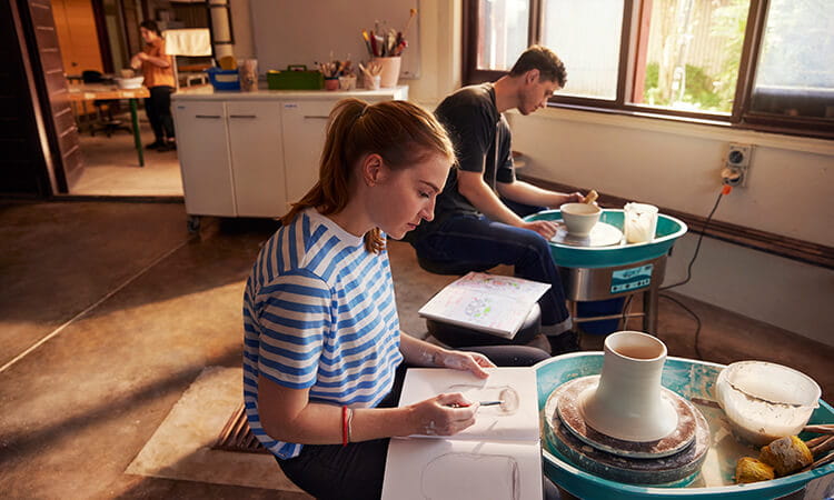 Two people work on pottery at wheels in a sunlit studio; a woman sketches in a notebook while a man shapes clay.