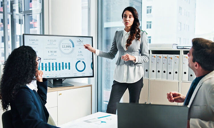 A businesswoman presents data charts on a screen to two colleagues in a modern office setting during a meeting.