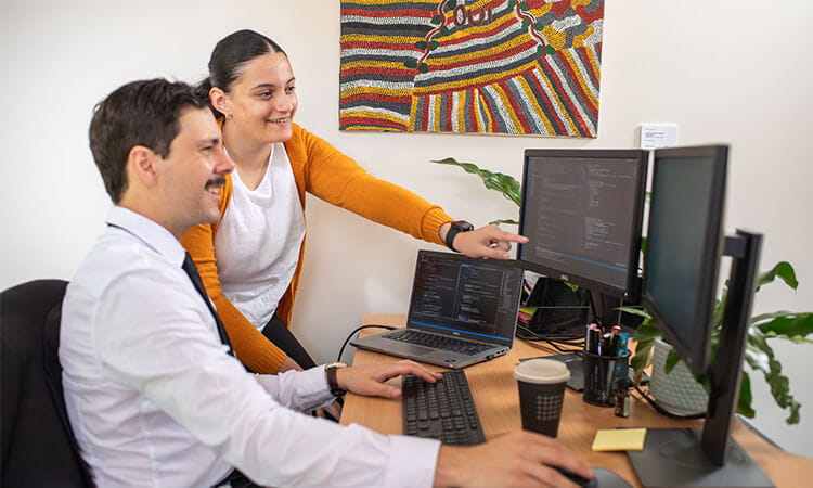 Two colleagues work at a desk with laptops and dual monitors; one is seated typing while the other stands, pointing at code on the screen. A colorful painting hangs on the wall behind them.