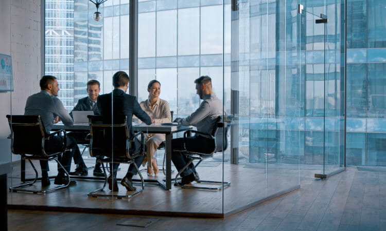 Five people in business attire sit around a conference table in a glass-walled office, engaged in a meeting with city buildings visible outside the windows.