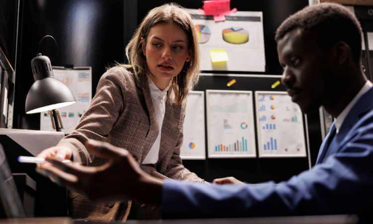 Two people in business attire review documents at a desk, with charts and graphs displayed on the wall behind them.