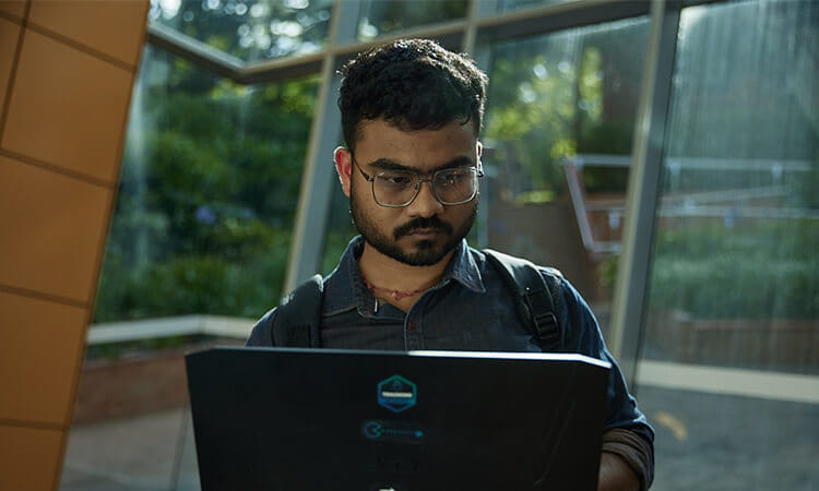 A man with glasses and a backpack looks at a laptop in a sunlit indoor space with large windows.