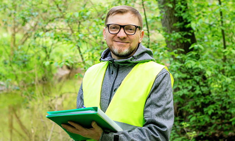 A person wearing glasses and a yellow safety vest holds folders and a pen, standing outdoors in a green, wooded area.