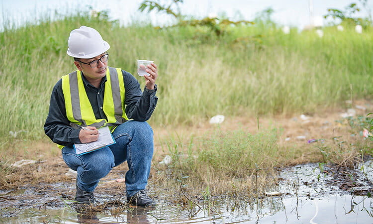 Man crouched down near water looking at water sample in jar.