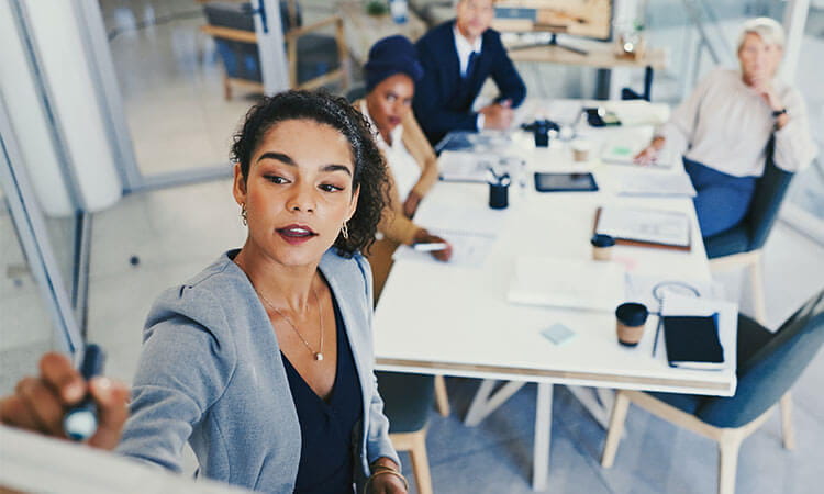 A woman writes on a whiteboard while three colleagues sit at a table with documents, laptops, and coffee cups in a modern office setting.