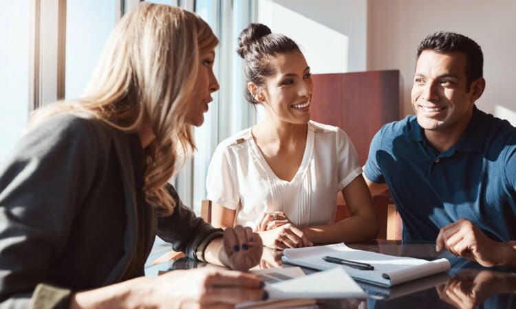 Three people sit at a table with documents and a pen, engaged in a discussion in a well-lit office setting.