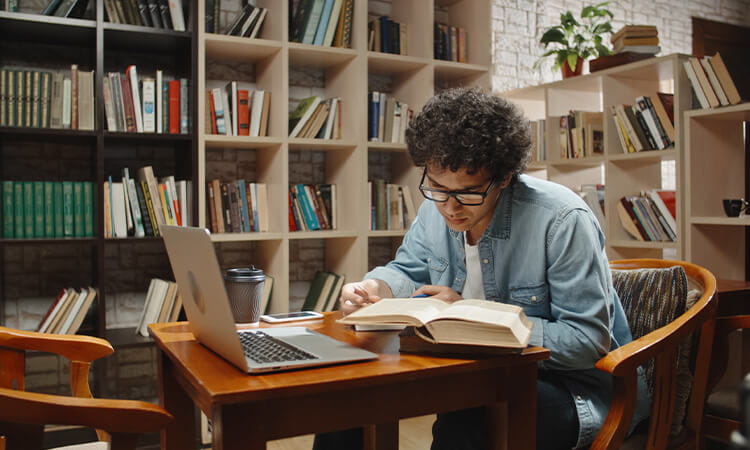 Man researching in library, reading book with laptop open. 