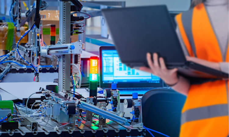Person in an orange safety vest holds a laptop near an automated assembly line with machinery, wires, and indicator lights in a factory setting.