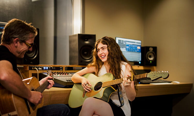 Two people sit in a recording studio playing acoustic guitars, with sound equipment and monitors visible in the background.
