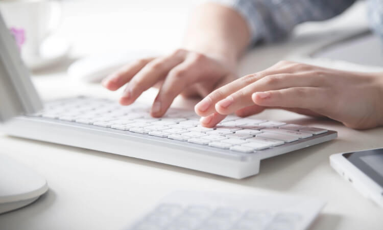 Close-up of a person typing on a white computer keyboard at a desk, with a smartphone and other office items visible nearby.