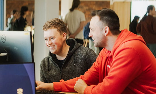 Two people sit at a desk, smiling and looking at a computer screen together. Other people are visible in the blurred background.